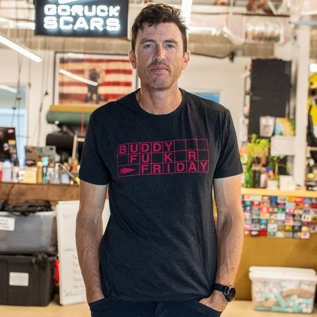 A man wearing a black t-shirt and shorts stands in an industrial workspace with a Buddy FCKR Friday sign behind him.