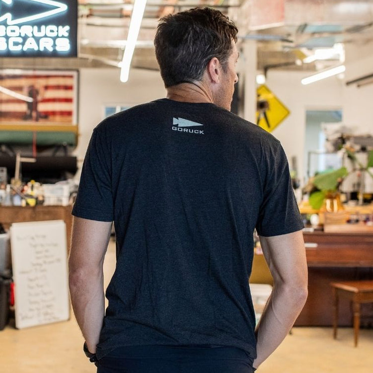 A man in a black “Easy Life Is Not For Us” t-shirt stands in an industrial-style room with a GORUCK SCARS sign and an American flag on the wall.