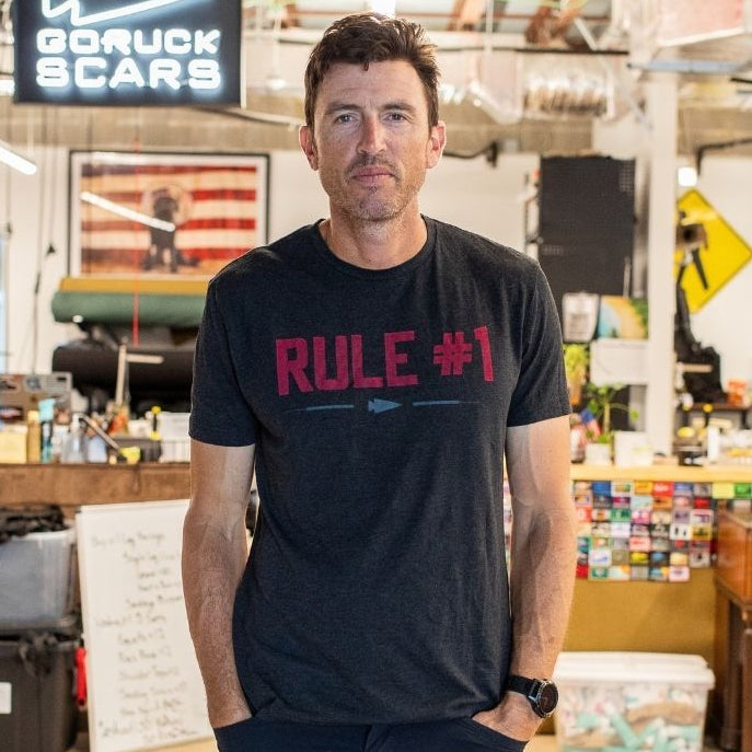 A man in a black “Rule 1” shirt stands in an industrial-style workspace with a GORUCK SCARS sign.