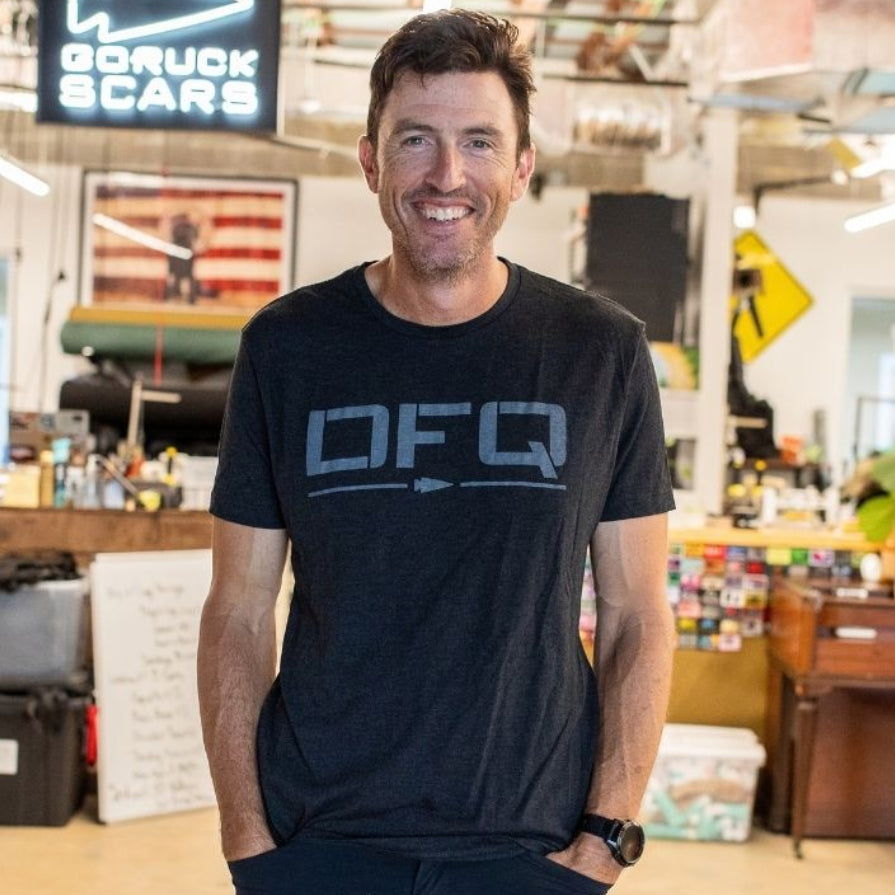 A man smiles while standing in a casual office with red beams, a piano, and a DFQ sign in the background.
