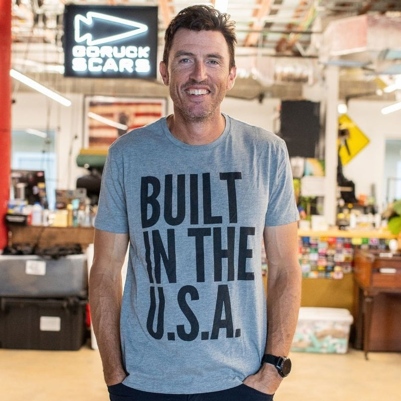A man smiles indoors while wearing the Graphic T-Shirt - Built in the USA (gray) in a modern workspace.