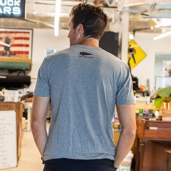 A man in a gray t-shirt stands in a workshop, facing away, with an “In GOD We Trust” sign visible ahead.