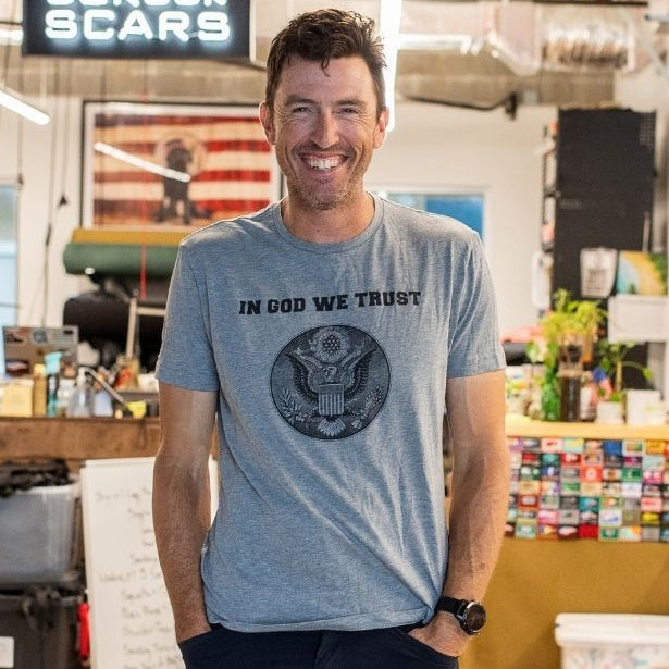 A man smiling in casual clothes stands in a workshop beneath a sign that reads “In GOD We Trust.”.