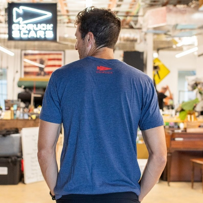 A man wearing the “America est. 1776” blue t-shirt stands indoors, facing away, with workshop equipment and signs in the background.