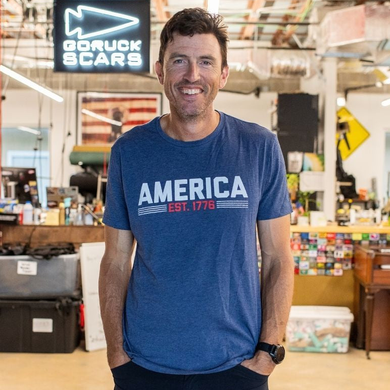 A man wearing the America est. 1776 shirt stands and smiles inside a casual office space.