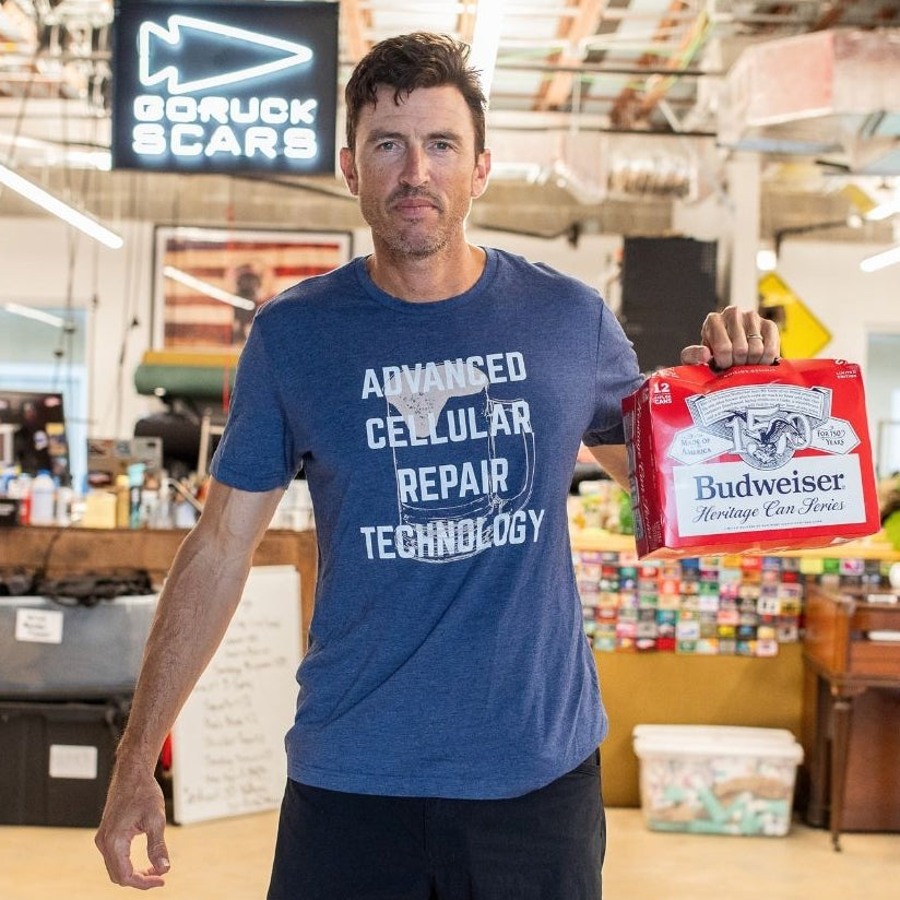 A man wearing a blue “ACRT” shirt holds a Budweiser box in a casual workspace.