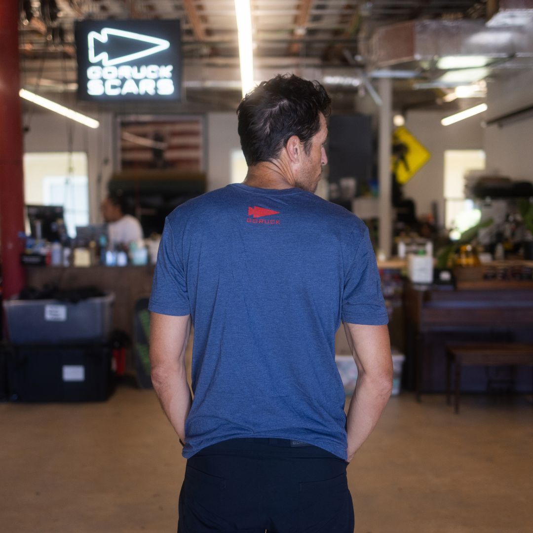 A man stands indoors with his back to the camera in a casual, cluttered workspace, wearing the DFQ TEE - TRI-BLENDX in blue. The motivational “Don’t F*ing Quit” message is displayed on the shirt, highlighting both its comfort and inspiring design.