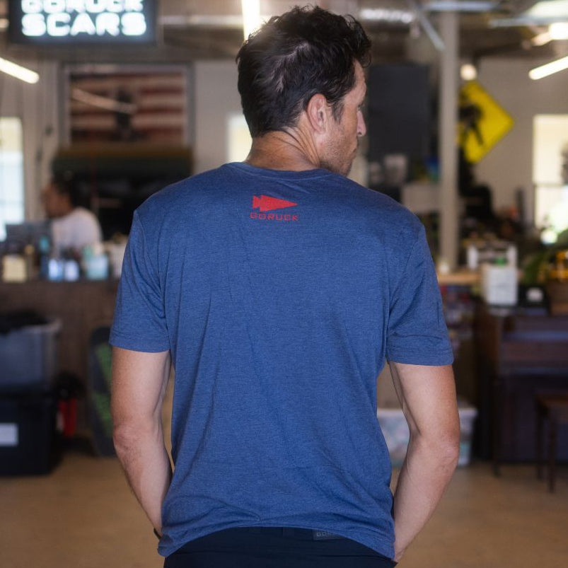 A man stands indoors with his back to the camera in a casual, cluttered workspace, wearing the DFQ TEE - TRI-BLENDX in blue. The motivational “Don’t F*ing Quit” message is displayed on the shirt, highlighting both its comfort and inspiring design.