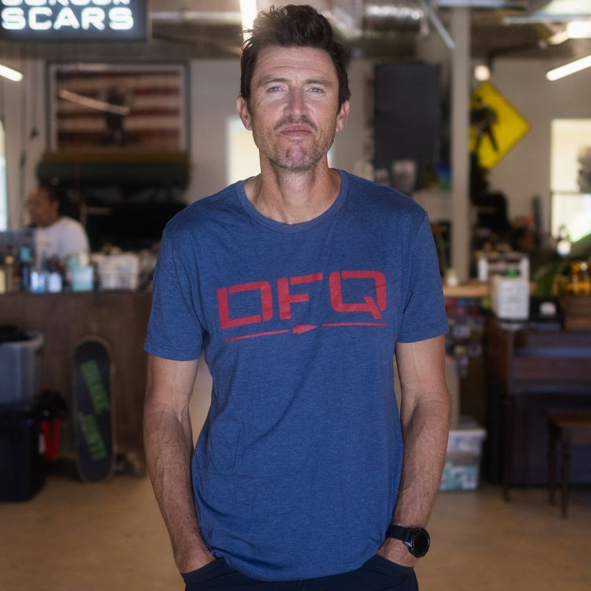 Wearing the DFQ TEE - TRI-BLENDX in blue, a man stands with his hands in his pockets inside a casual, industrial-style workspace. The bold “Don’t F*ing Quit” message on his shirt adds determination to the modern vibe.