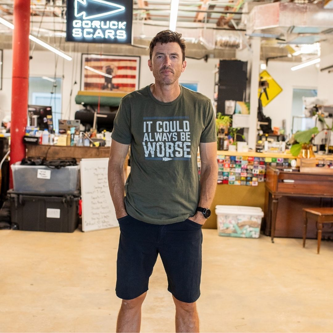 A man in casual clothes stands in a cluttered workshop, wearing the “It Could Always Be Worse” T-shirt—a premium tri-blend shirt printed in the USA.