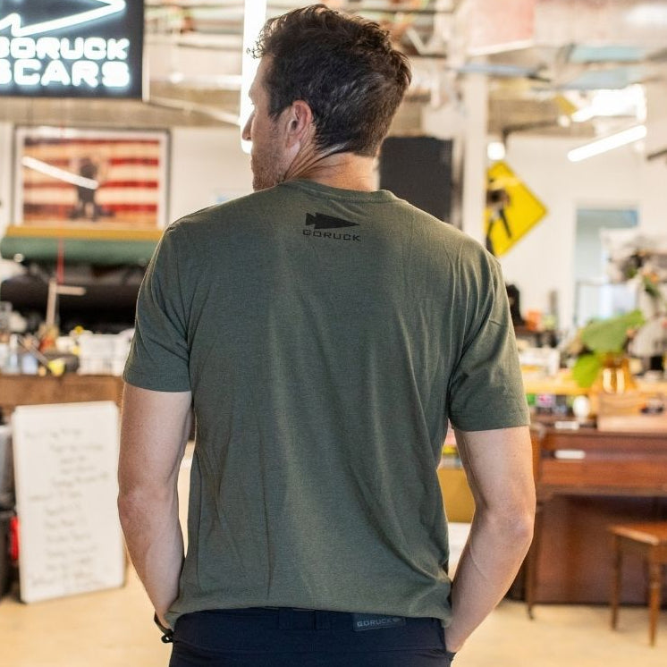 A man in a green T-shirt stands indoors facing away, with a DFQ sign and an American flag in the background.