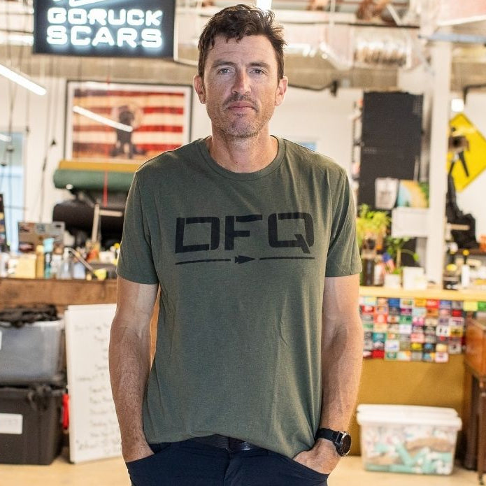 A man in a green T-shirt stands in an open office near a piano, facing the camera with his hands in his pockets, wearing DFQ.