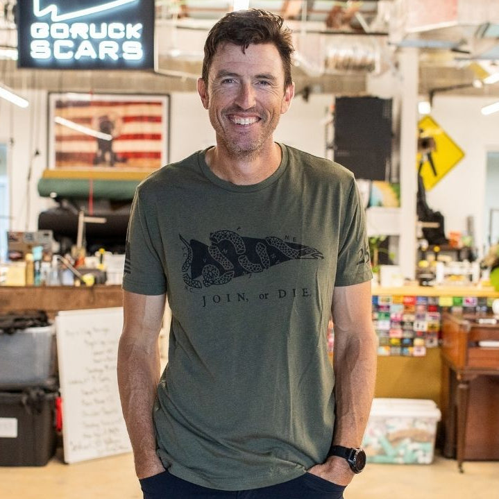 A man wearing the Join or Die Tee - Tri-Blend stands smiling in a workshop with red pipes and a “GORUCK SCARS” sign behind him, evoking the spirit of colonial unity first championed by Ben Franklin through his famous “Join or Die” cartoon.