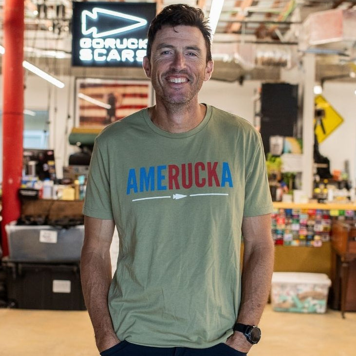 A man smiles in a workshop, wearing an “AmeRUCKa” shirt with his hands in his pockets; a busy workspace is visible behind him.