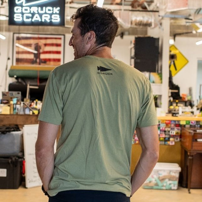 A man in a green T-shirt stands in a cluttered office beneath a sign reading “DFQ.”.
