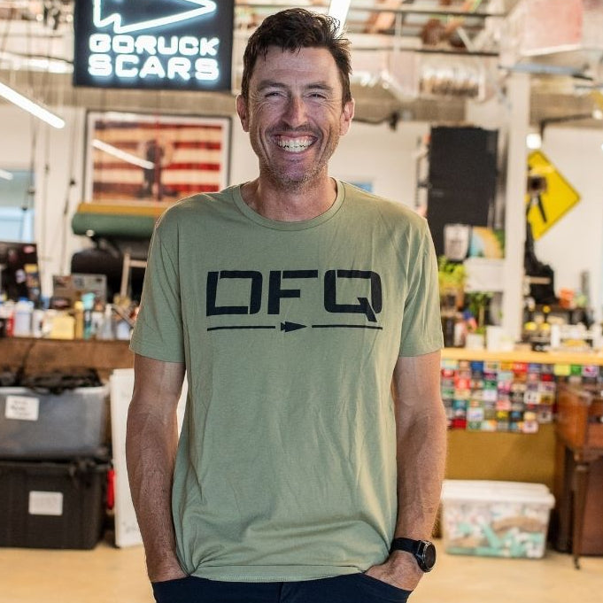 A man wearing a DFQ casual t-shirt smiles, standing in a bright, cluttered workspace with industrial decor.