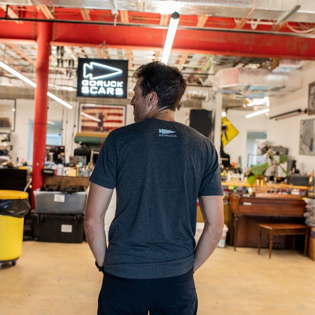 A man wearing the “It Could Always Be Worse” premium tri-blend t-shirt stands in a workspace with red beams, facing away from the camera.