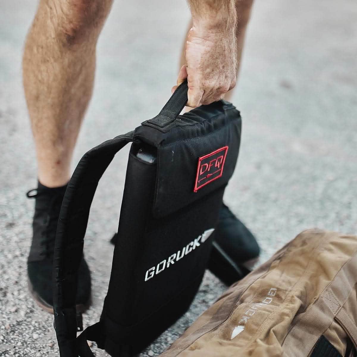 Person lifting a GORUCK black ruck plate with DFQ patch next to a tan GORUCK bag on pavement