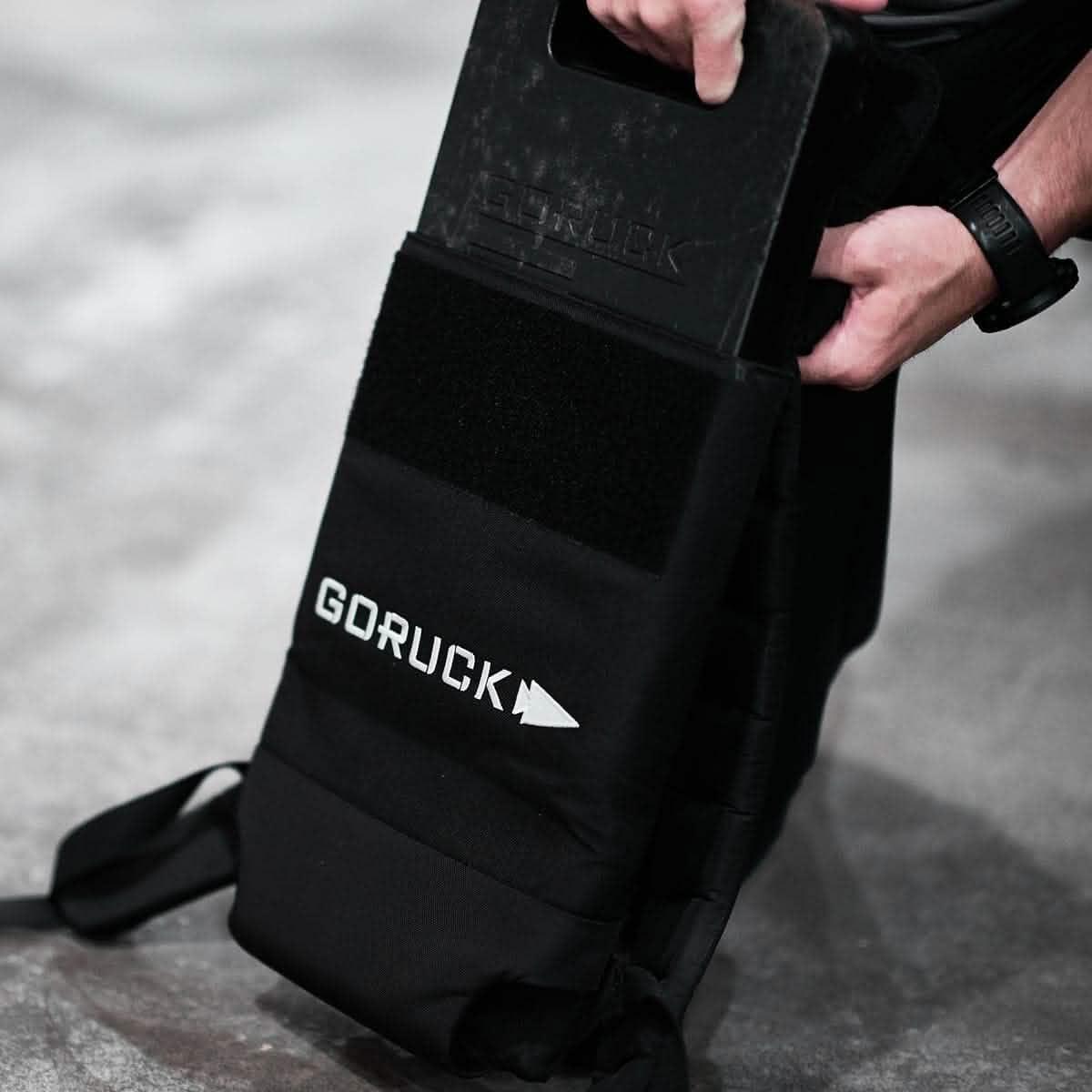 Hands inserting a black GORUCK 45lb ruck plate into a GORUCK backpack on concrete floor