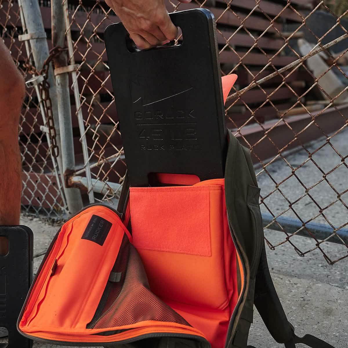 Hand placing a black 45LB GORUCK ruck plate into an open olive green and bright orange rucksack next to a chain link fence