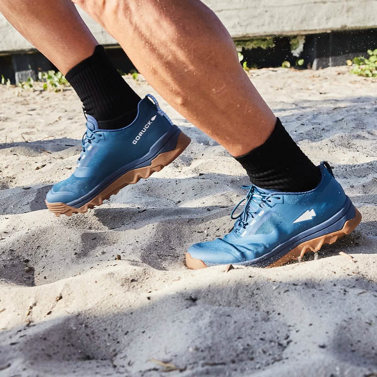 Close-up of a person running on a sandy surface wearing GORUCK's Mackall - Legion Blue + Gum trail shoes, paired with black socks and shorts. The image captures the dynamic motion typical in Special Forces training, with sand displaced by each powerful step.
