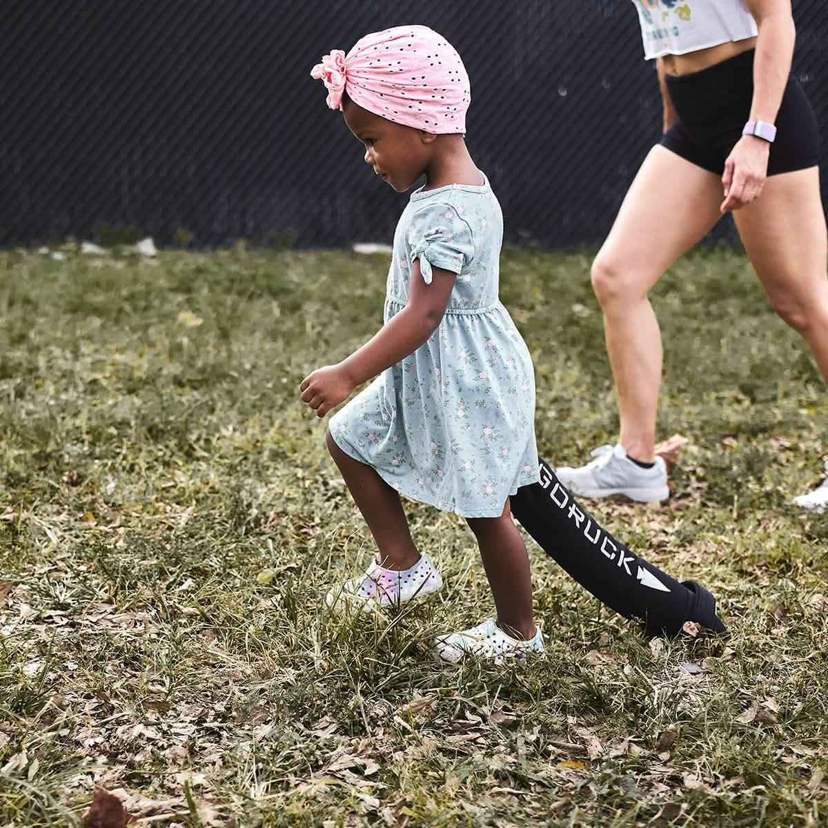 Child in a pink turban and floral dress walking outdoors with an adult wearing GORUCK gear on grass