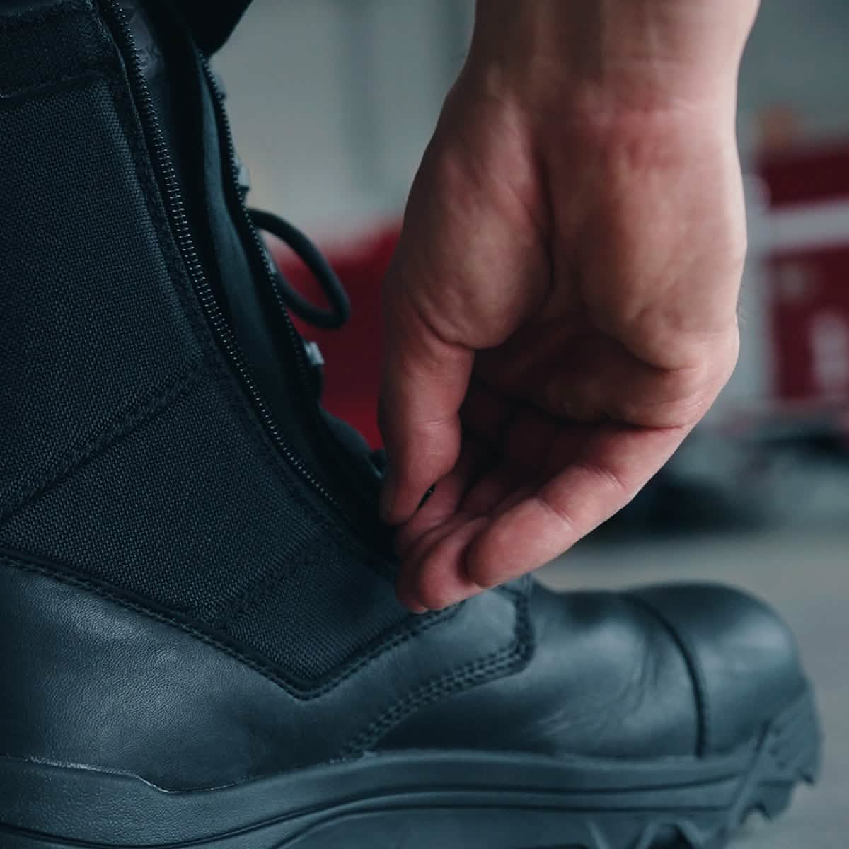 A close-up shows someone zipping up a GORUCK MACV-2 Safety Boot - High Top. Their hand pulls the zipper upwards, showcasing the rugged sole that meets Special Forces standards. In the slightly blurred background, there's a palpable sense of safety and durability in every step.