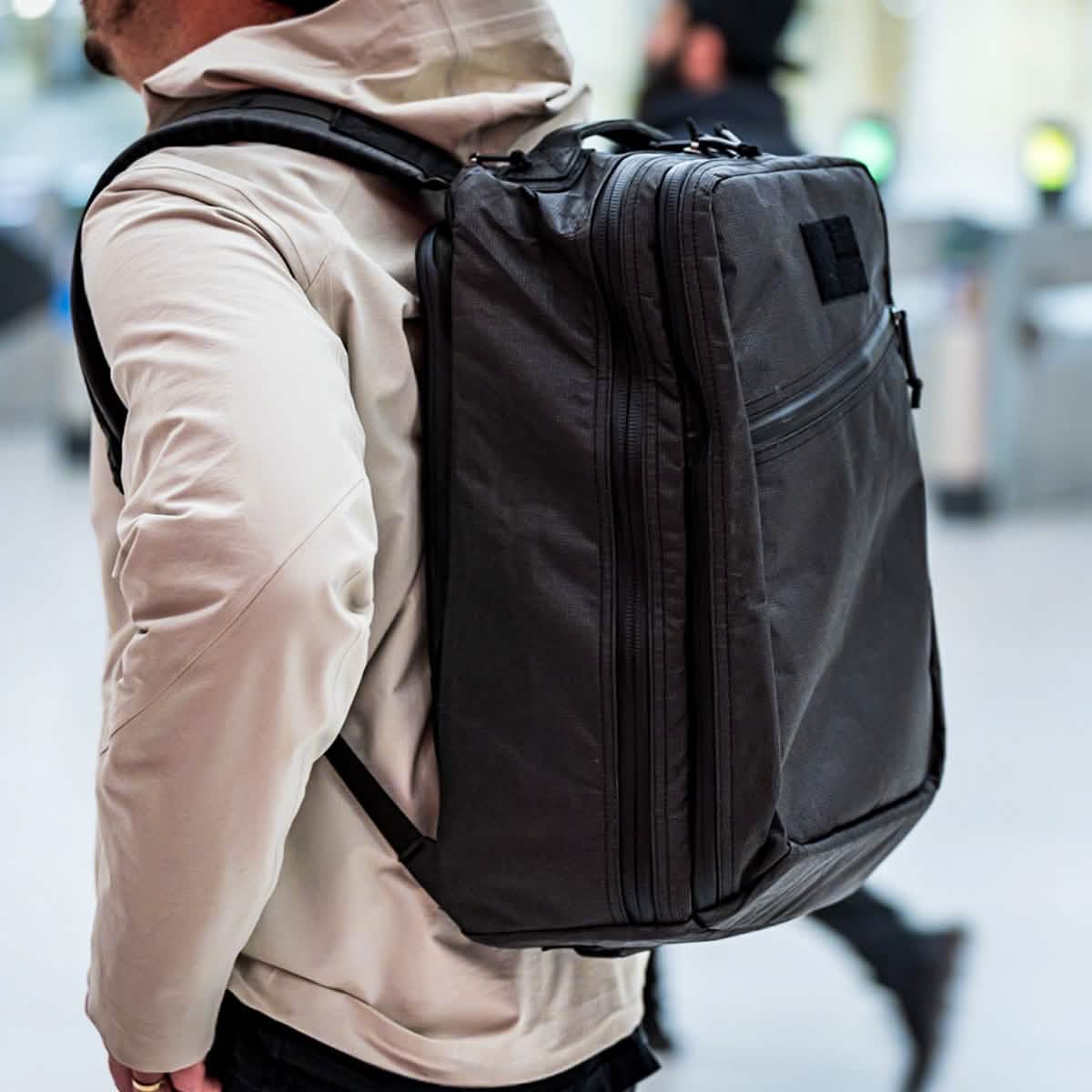 A person wearing a beige jacket carries the GR2 USA x Carryology - Ultra Black backpack, crafted from UHMWPE fabric, in an indoor setting.