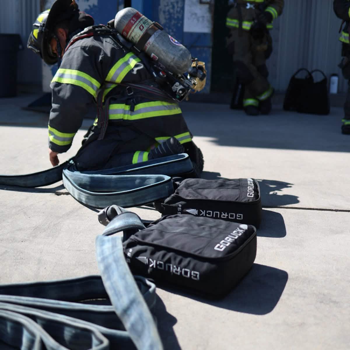 A firefighter in full gear kneels on the ground, showcasing core strength as he manages a fire hose. In the foreground sit two black GORUCK Sand Jerry Cans, crafted from Special Forces Grade materials, with additional equipment visible in the background, suggesting preparation or training.