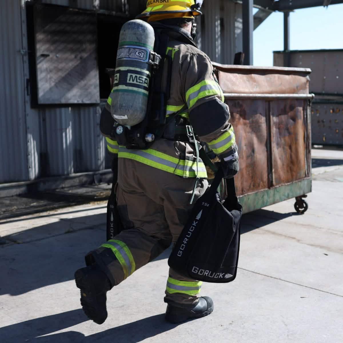firefighter in full gear carrying black GORUCK sandbags during training near industrial building