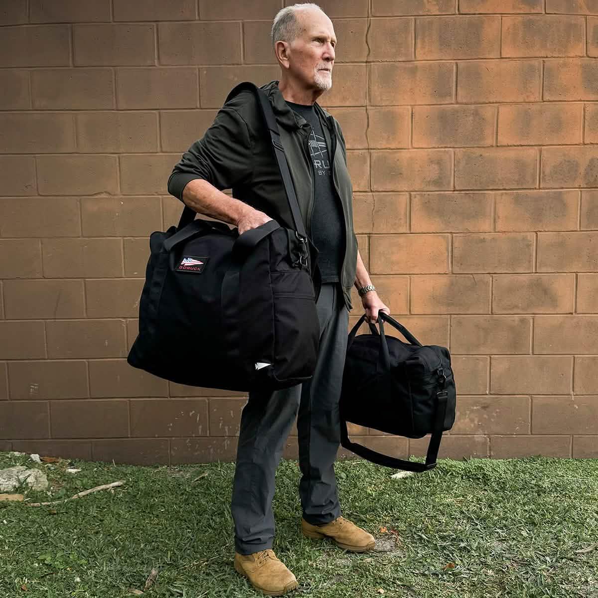 Man holding two GORUCK rucking bags outdoors against a brick wall, showcasing durable gear