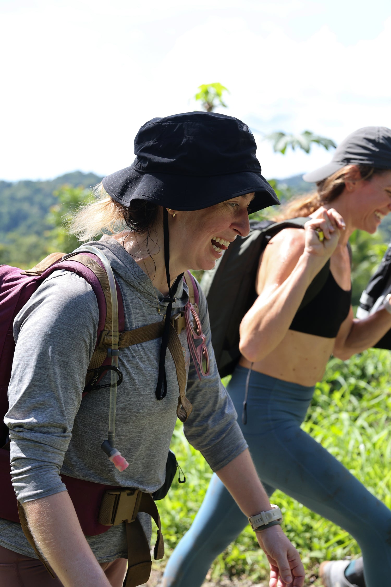 Two women in activewear and backpacks smiling while hiking outdoors on a sunny day.