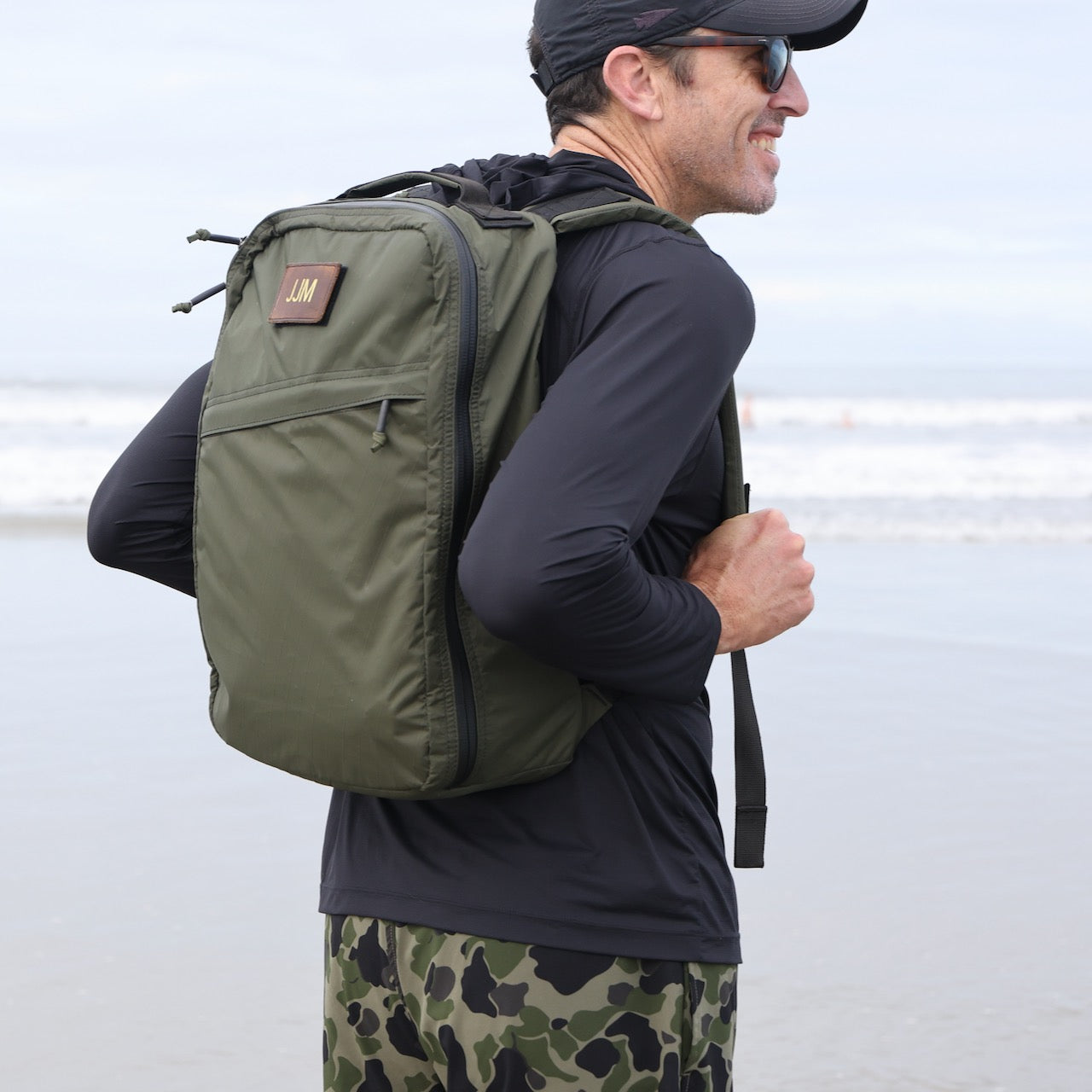 Man wearing a green backpack featuring the Patch - Custom Leather Monogram, black shirt, and camo shorts stands on a beach smiling.