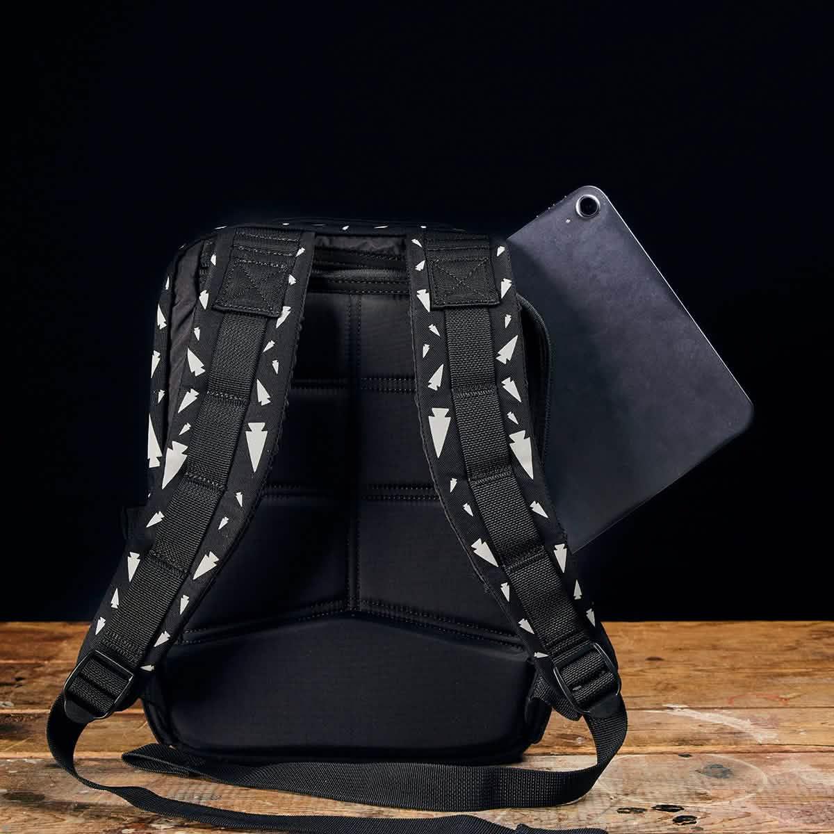 The REFLECTIVE KR1, a compact black school backpack with patterned straps and a tablet partly tucked in the back pocket, rests on a wooden surface.