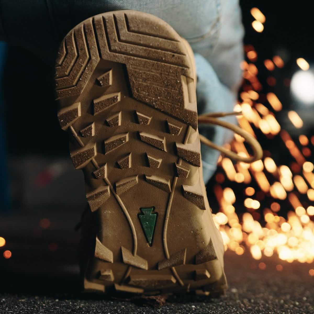 A close-up of a person stepping forward in the GORUCK MACV-2 Safety Boot - Mid Top, showcasing its rugged sole with prominent tread patterns. Sparks fly in the background, enhancing the dynamic and industrial atmosphere. The jeans above the boot contribute to the sense of action, aligning with Special Forces standards for durability and toughness.