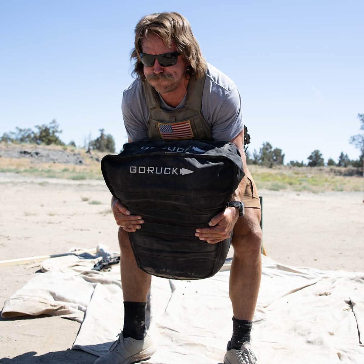 Man wearing tactical gear lifting a black GORUCK weighted bag outdoors in a dry, desert setting