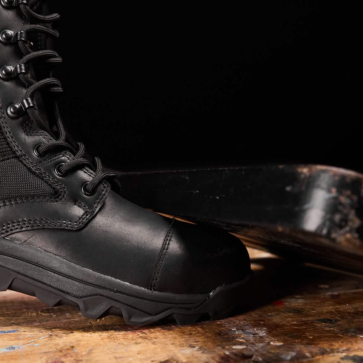Close-up of a MACV-2 Safety Boot - High Top by GORUCK in black, featuring rugged soles that meet Special Forces standards, resting on a wooden surface against a dark background.