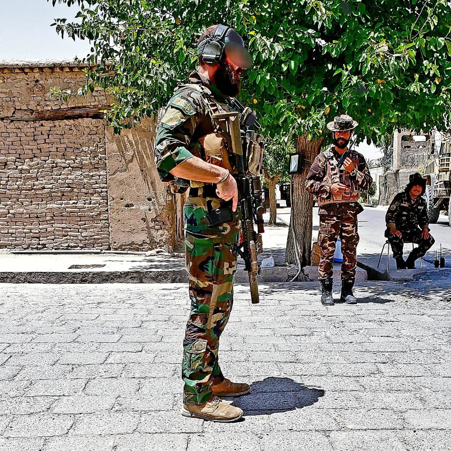 A soldier in camouflage, sporting a beard and helmet, stands firmly with a rifle on a paved area. His feet are clad in GORUCK's MACV-1 Hi-Speed - High Top boots. Behind him, two fellow soldiers rest under a tree by a brick wall, enjoying the sunlit setting.