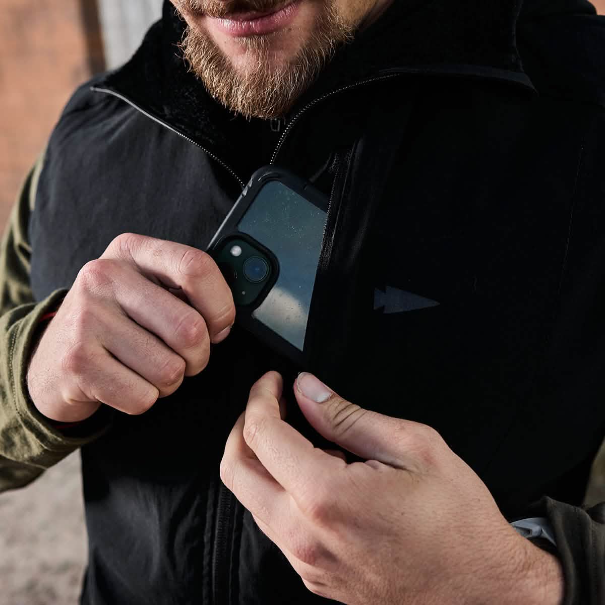 A person is slipping a smartphone with a black case into the chest pocket of their black jacket, echoing the style of GORUCK's Vest of Power - ToughDry® + Fleece. Only their hands and part of their face are visible, revealing a green long-sleeved shirt underneath, suggesting an environmentally conscious choice.