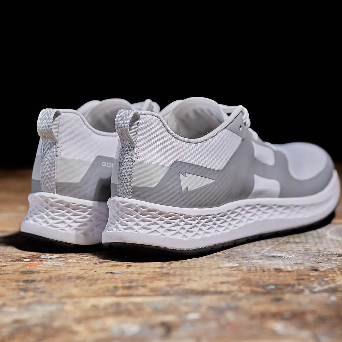 A pair of Men's Rough Runner - White + Speed Grey athletic shoes by GORUCK, featuring textured soles, are displayed on a wooden surface. These high mileage running shoes showcase a geometric arrow design and the Gradient Density™ EVA Midsole, with their heels facing the camera against a dark background.