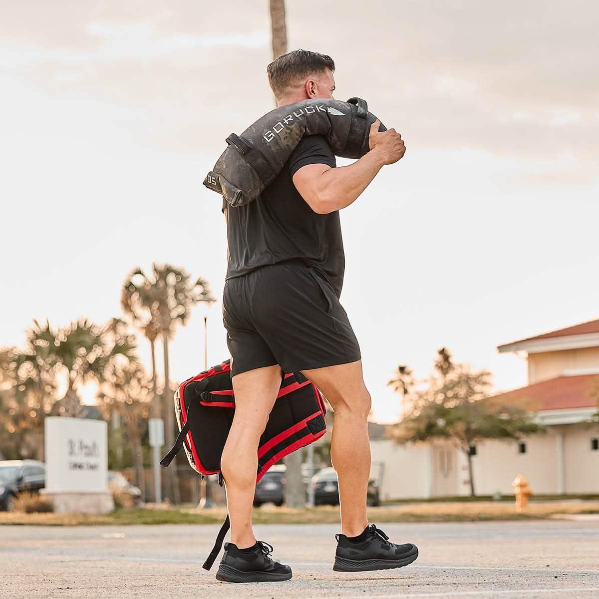 Athletic man carrying a black GORUCK weighted sandbag on shoulder outdoors, wearing black workout gear