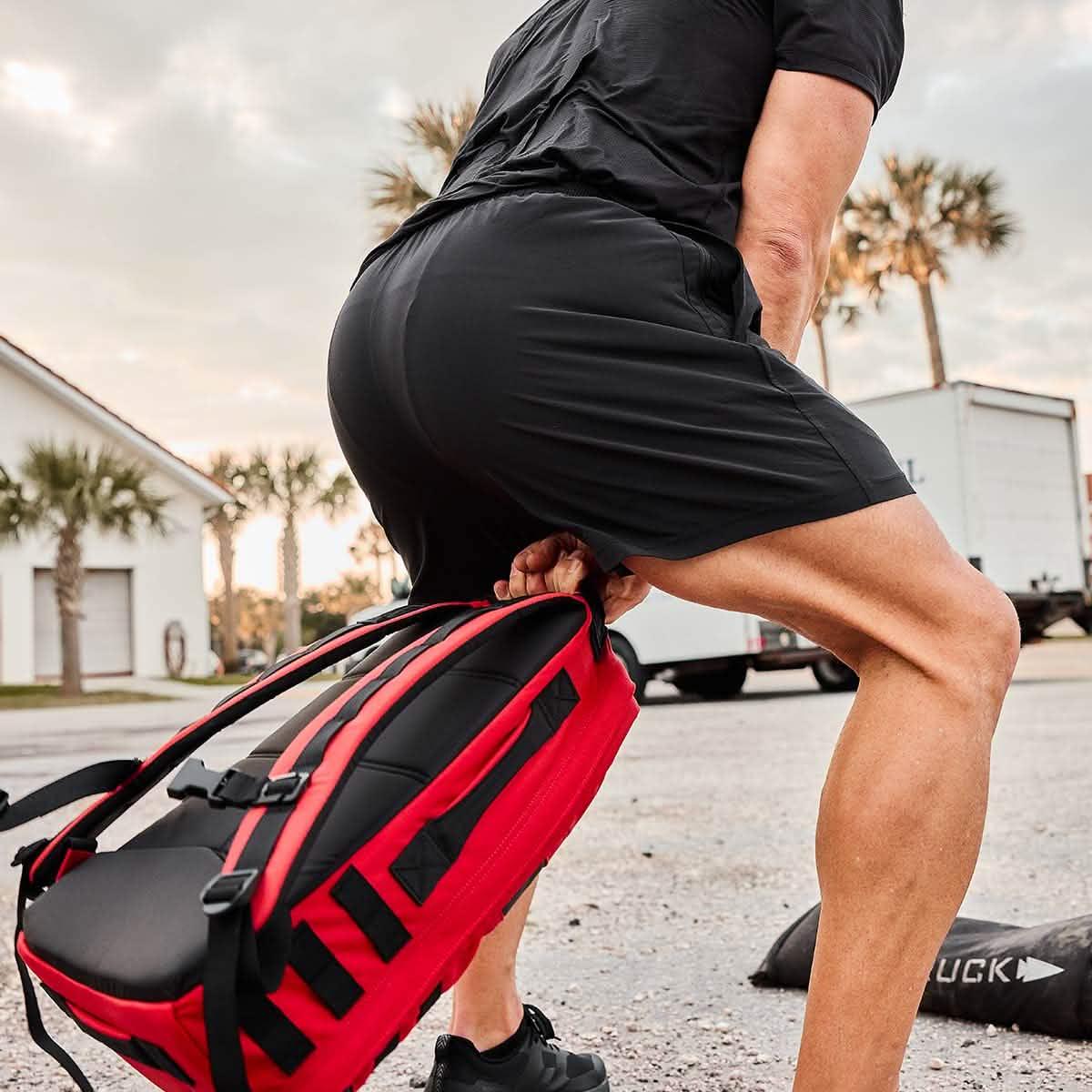 Person lifting a red and black tactical rucksack outdoors, wearing black athletic shorts and shirt