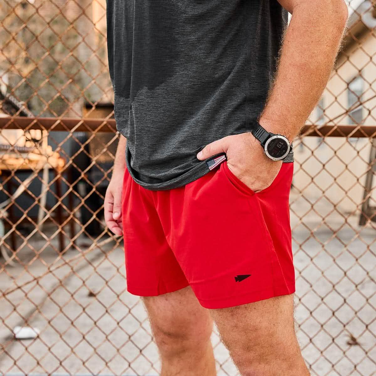 Man wearing GORUCK red rucking shorts and black shirt by a chain-link fence outdoors