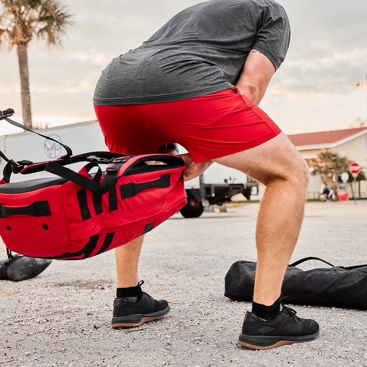 Man lifting a rugged red GORUCK rucksack outdoors, wearing red shorts and black shoes