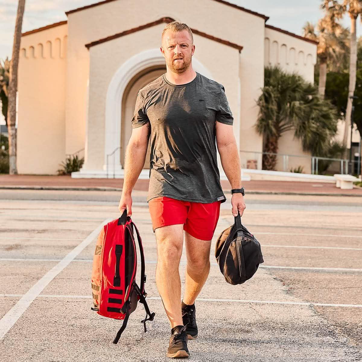Man in athletic gear carrying a GORUCK red rucksack and black duffel bag outdoors