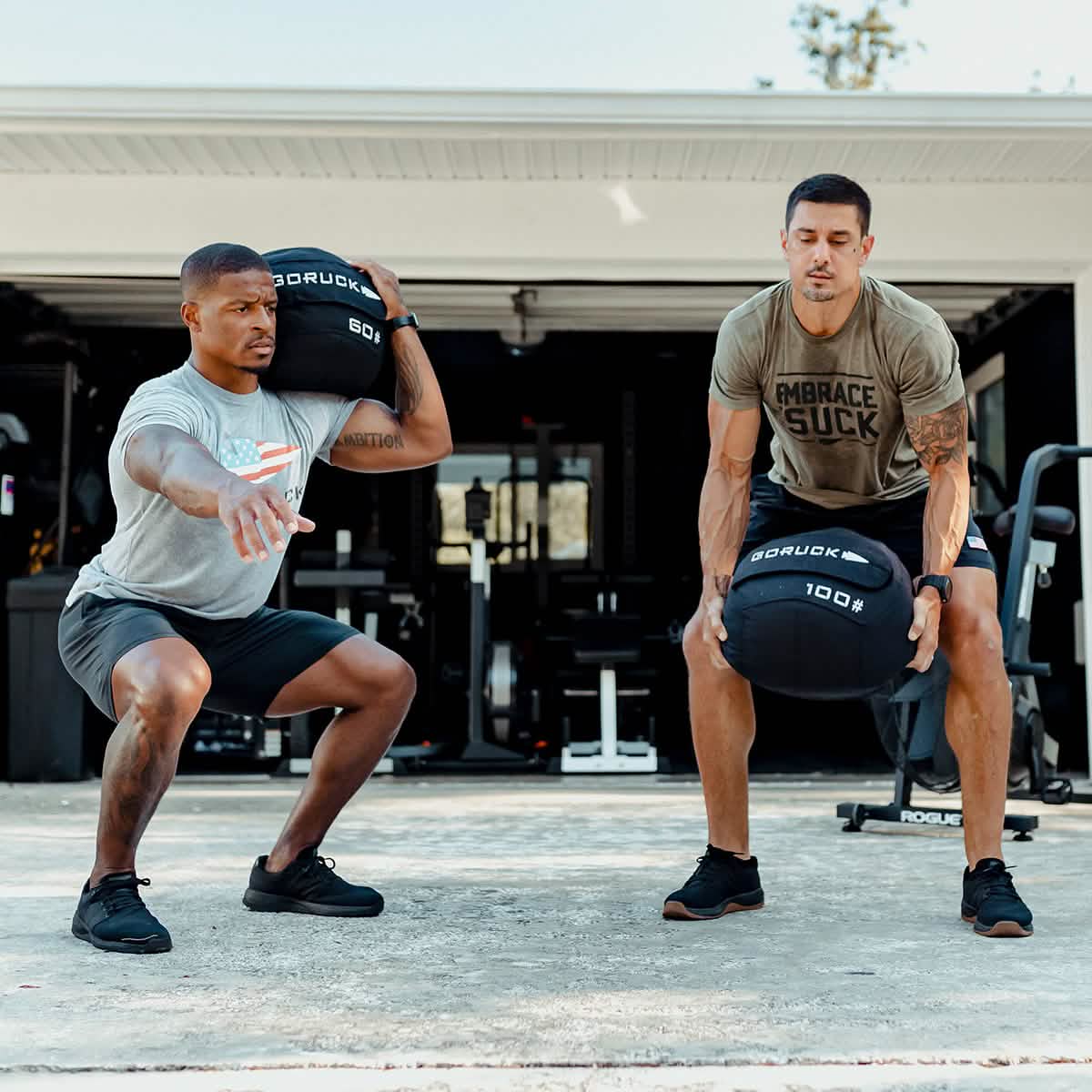 Two men are exercising outdoors in front of a home gym, each holding a heavy black GORUCK Sand Medicine Ball. The man on the left is squatting, while the man on the right stands focused, enhancing his core stability in athletic clothing.
