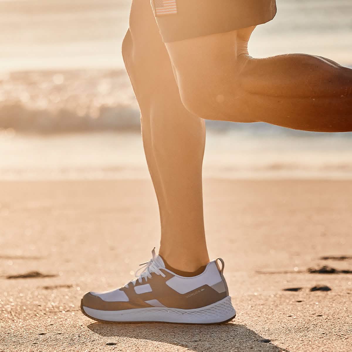 Close-up of a person jogging on a sunny beach, legs in motion and wearing GORUCK's Men's Rough Runner in White + Speed Grey. The ocean serves as a scenic backdrop, complementing the sturdy footwear designed for optimal performance.