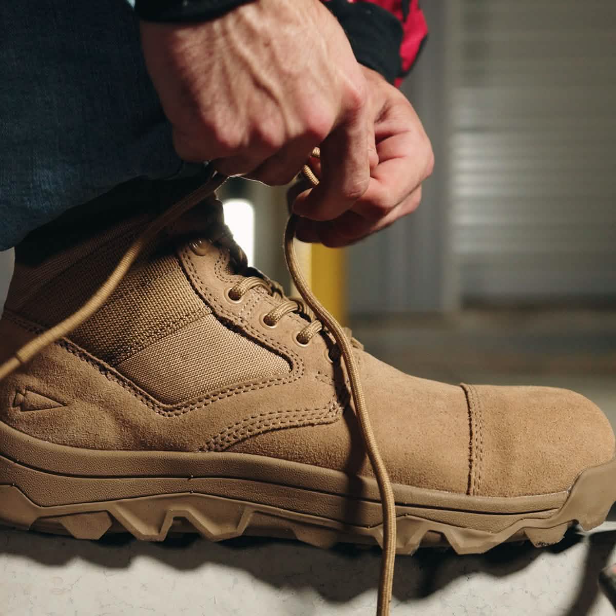 A close-up of someone fastening the laces on a tan MACV-2 Safety Boot - Mid Top from GORUCK, designed to meet Special Forces Standards. Ideal for demanding tasks, the boots are worn over denim jeans while the background remains softly blurred, suggesting an indoor environment.
