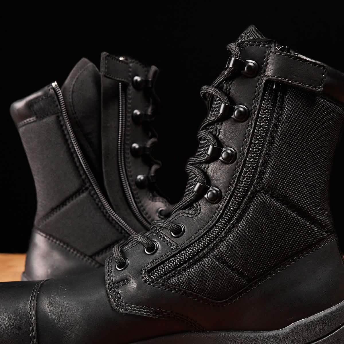 A close-up showcases the GORUCK MACV-2 Safety Boots - High Top on a wooden surface, engineered to Special Forces standards. These black boots include laces, eyelets, zippers, and padded sections for robust construction. Against a solid black background, the high-top design stands out as the ultimate work boot.