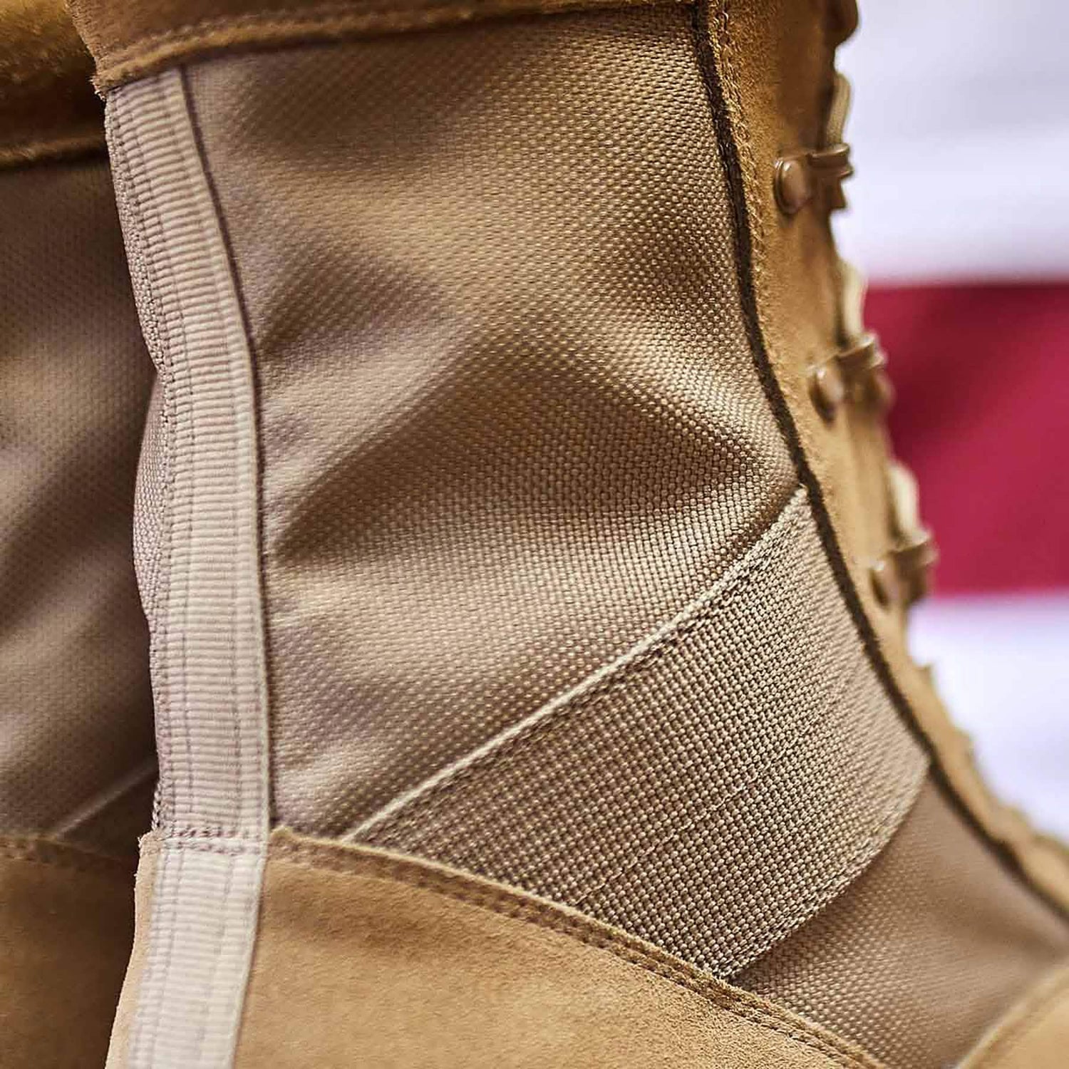 Close-up of the MACV-1 Hi-Speed - High Top boot from GORUCK, showcasing its detailed stitching and rugged fabric while meeting AR670-1 compliance standards. The lightweight rucking boot is laced up tightly, emphasizing its durable build. A blurred red and white background adds depth to the image, enhancing its military-ready appeal.